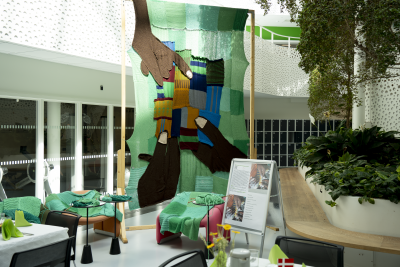 Et gigantisk strikkecollage, skabt i et samarbejde mellem strikke-entusiaster med synshandicap i henholdsvis Rwanda og Danmark og en billedkunstner pryder billedet med sin grønne farve og tre mørke hænder, der mødes på midten. 