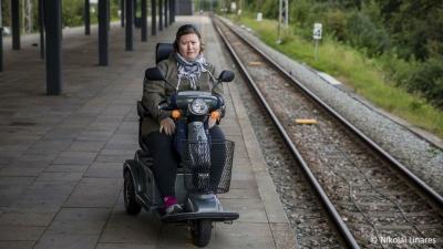 Kvinde sidder i el-scooter på en togstation. Hun sidder med front til kameraet og til højre for hende ser man togskinnerne