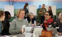 To kvinder fra Foreningen Danske Døvblinde sidder om et bord og deltager smilende i en samtale. I baggrunden ses snakkende mennesker foran flipcharts.