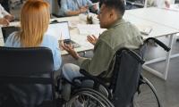 Mand i kørestol sidder i et mødelokale om et bord med sine kollegaer. De snakker om noget arbejdsrelateret