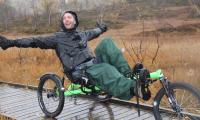 Mand i regntøj kører trehjulet cykel på en træbelagt sti i naturen